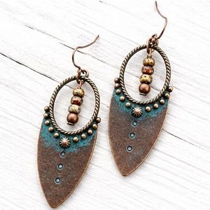 May include: A pair of drop earrings featuring a teardrop-shaped copper-toned metal base with a textured surface and a teal patina finish. Each earring includes a circular twisted metal frame, three small beads, and a curved ear wire hook.