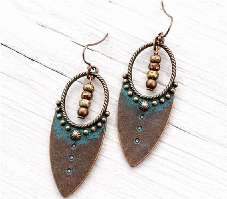 May include: A pair of drop earrings featuring a teardrop-shaped copper-toned metal base with a textured surface and a teal patina finish. Each earring includes a circular twisted metal frame, three small beads, and a curved ear wire hook.