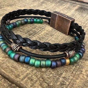 Puede incluir: Un brazalete de cuero negro con un dise&ntilde;o trenzado y un cierre magn&eacute;tico. El brazalete tiene tres filas de cuentas iridiscentes verdes, azules y doradas.