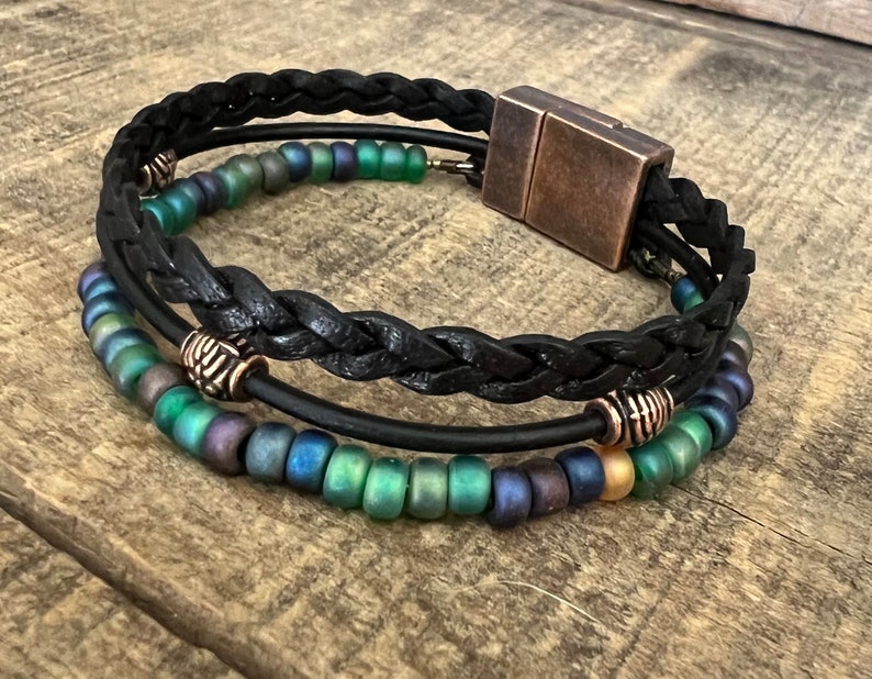 Puede incluir: Un brazalete de cuero negro con un dise&ntilde;o trenzado y un cierre magn&eacute;tico. El brazalete tiene tres filas de cuentas iridiscentes verdes, azules y doradas.