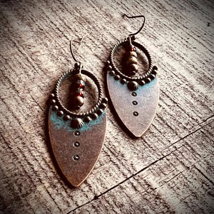 May include: A pair of drop earrings featuring a teardrop-shaped copper-toned metal base with a teal patina accent. Each earring includes a circular twisted wire frame, a vertical row of three small dark beads, and three punched circular holes.