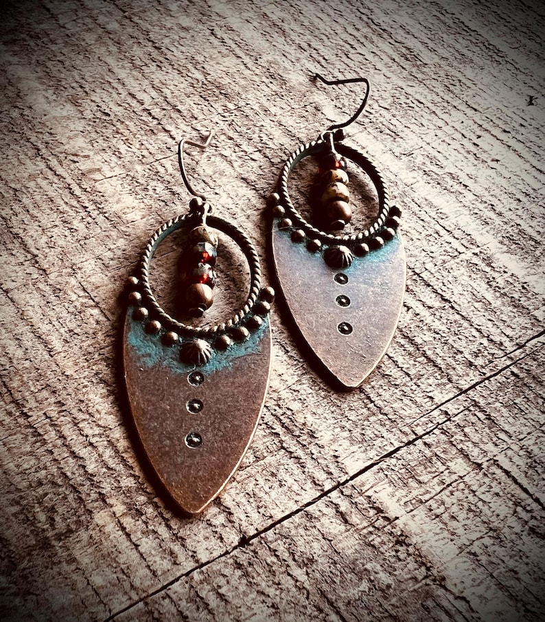 May include: A pair of drop earrings featuring a teardrop-shaped copper-toned metal base with a teal patina accent. Each earring includes a circular twisted wire frame, a vertical row of three small dark beads, and three punched circular holes.