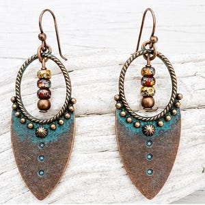 May include: A pair of dangle earrings featuring a copper-toned teardrop-shaped base with a teal patina finish. Each earring includes a twisted circular frame, a small sunburst detail, and a vertical row of three beads in mottled earth tones.