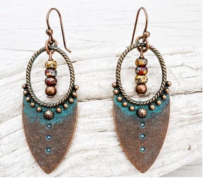 May include: A pair of dangle earrings featuring a copper-toned teardrop-shaped base with a teal patina finish. Each earring includes a twisted circular frame, a small sunburst detail, and a vertical row of three beads in mottled earth tones.