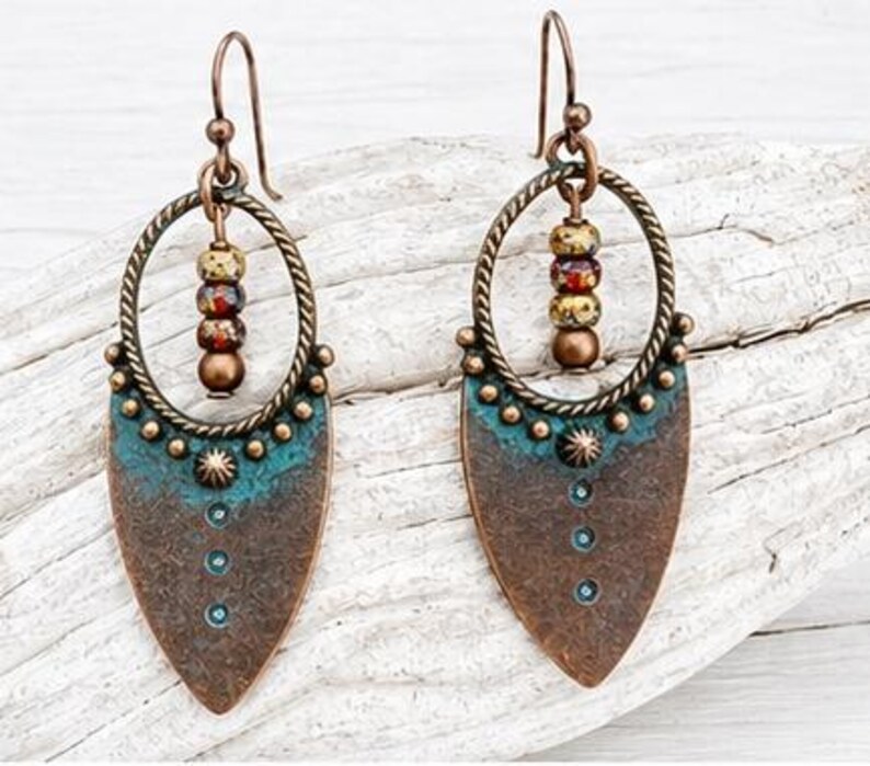 May include: A pair of dangle earrings featuring a copper-toned teardrop-shaped pendant with a teal patina finish, accented by small metal studs and three vertical dots. A twisted metal hoop hangs above, holding three small multicolored beads.