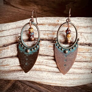 May include: A pair of dangle earrings featuring a teardrop-shaped copper base with a dark brown finish and a teal patina accent. Each earring includes a circular twisted wire frame, three small stacked beads in red and tan, and metal studs.
