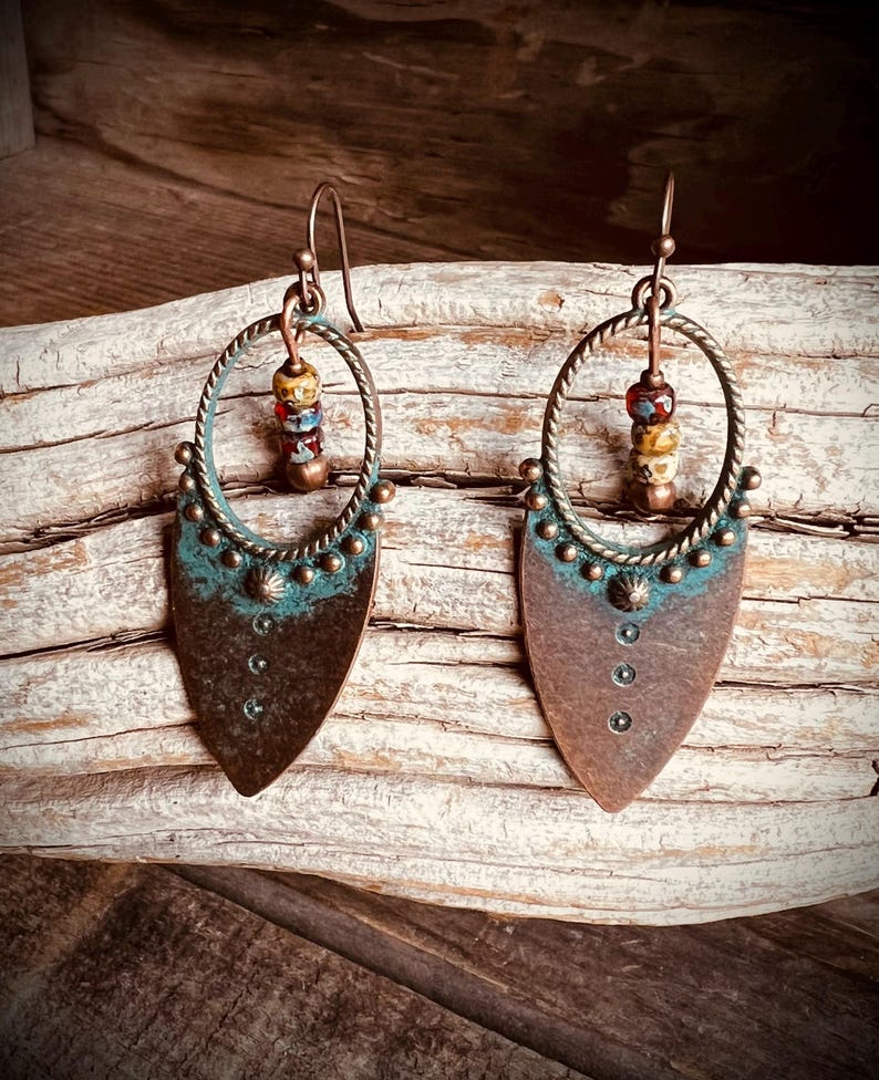 May include: A pair of dangle earrings featuring a teardrop-shaped copper base with a dark brown finish and a teal patina accent. Each earring includes a circular twisted wire frame, three small stacked beads in red and tan, and metal studs.