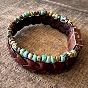 Braided brown leather bracelet with turquoise and multicolored beads. The bracelet features a copper-colored clasp. The beads are a mix of blue, yellow, and brown. The bracelet is shown on a wooden surface.