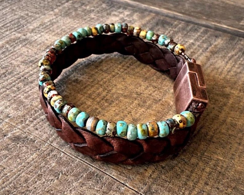 Braided brown leather bracelet with turquoise and multicolored beads. The bracelet features a copper-colored clasp. The beads are a mix of blue, yellow, and brown. The bracelet is shown on a wooden surface.