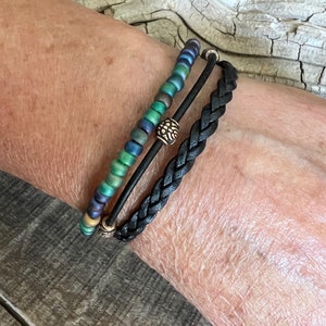 Puede incluir: Un conjunto de tres pulseras, una pulsera de cuero trenzado negro y dos pulseras de cuentas con detalles dorados. Las pulseras de cuentas presentan cuentas iridiscentes verdes, azules y doradas.