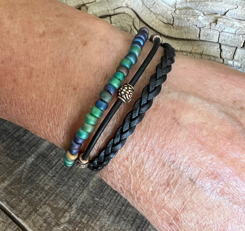 Puede incluir: Un conjunto de tres pulseras, una pulsera de cuero trenzado negro y dos pulseras de cuentas con detalles dorados. Las pulseras de cuentas presentan cuentas iridiscentes verdes, azules y doradas.