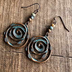 May include: A pair of copper-coloured earrings featuring a spiral design, highlighted with turquoise blue and small beads. The earrings have a rustic, vintage aesthetic, with a hook closure. The background is a wooden surface.
