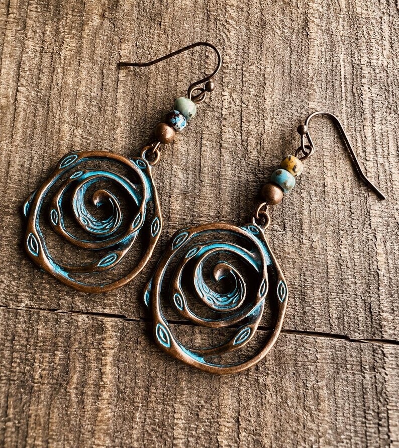 May include: A pair of copper-coloured earrings featuring a spiral design, highlighted with turquoise blue and small beads. The earrings have a rustic, vintage aesthetic, with a hook closure. The background is a wooden surface.
