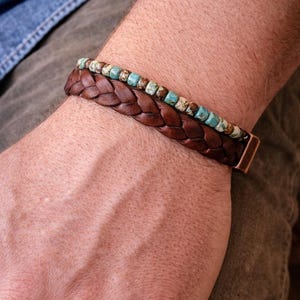 A close-up of two bracelets. The bottom bracelet is made of braided brown leather. The top bracelet has small turquoise and brown beads. The bracelets are on a person's wrist.