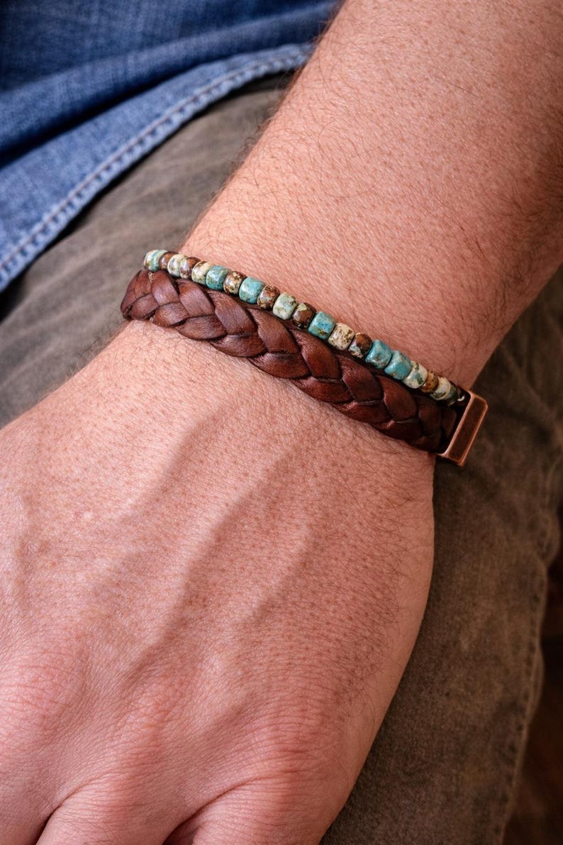A close-up of two bracelets. The bottom bracelet is made of braided brown leather. The top bracelet has small turquoise and brown beads. The bracelets are on a person's wrist.