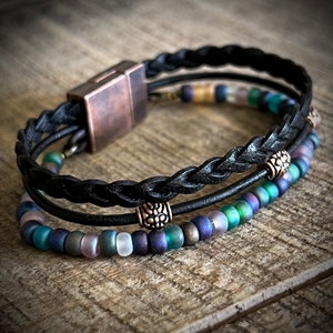 May include: A black leather bracelet with a braided design and a magnetic clasp. The bracelet is adorned with iridescent blue, green, and purple beads and silver accents.
