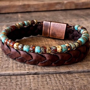 Braided brown leather bracelet with a turquoise and brown beaded accent. The bracelet features a copper-colored magnetic clasp. The design is rustic and casual, suitable for everyday wear.