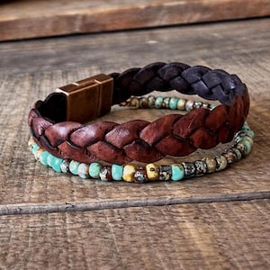 Three bracelets on a wooden surface. The top bracelet is dark brown braided leather with a gold clasp. The middle bracelet is a lighter brown braided leather. The bottom bracelet is made of turquoise and brown beads.