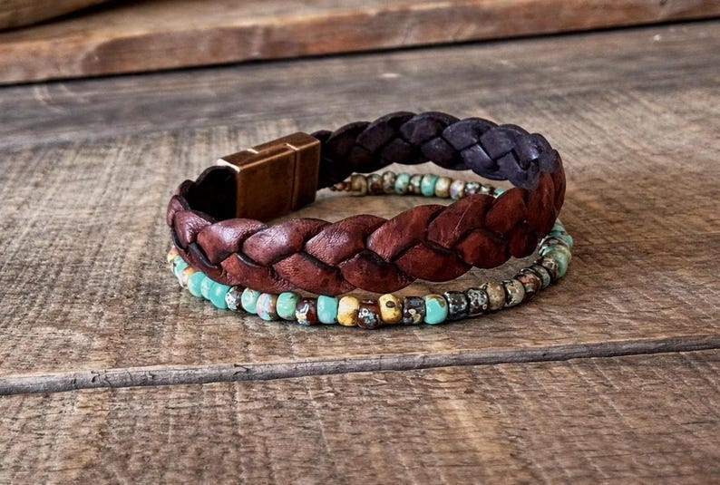 Three bracelets on a wooden surface. The top bracelet is dark brown braided leather with a gold clasp. The middle bracelet is a lighter brown braided leather. The bottom bracelet is made of turquoise and brown beads.