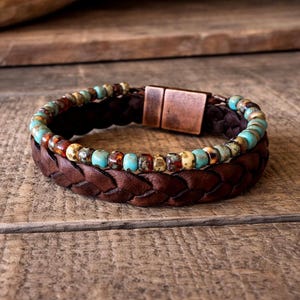 A two-layer bracelet with a braided brown leather band and a turquoise and brown beaded strand. The bracelet features a copper-colored magnetic clasp. The jewelry is displayed on a wooden surface.