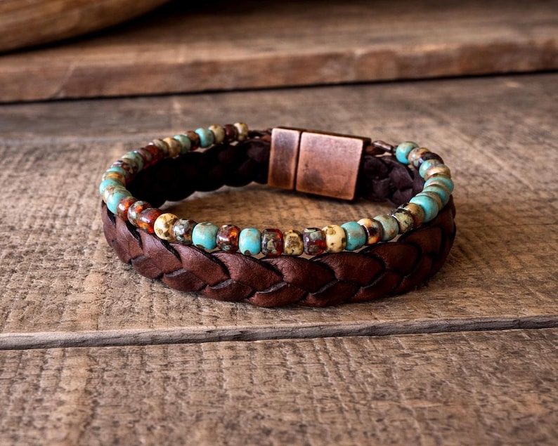 A two-layer bracelet with a braided brown leather band and a turquoise and brown beaded strand. The bracelet features a copper-colored magnetic clasp. The jewelry is displayed on a wooden surface.