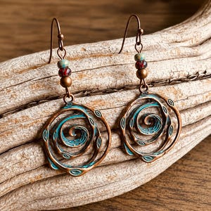 May include: Copper-coloured earrings featuring a spiral design with turquoise detailing. The earrings have a hook fastening and are decorated with small beads in red, green, and brown. The earrings are displayed on a piece of driftwood.