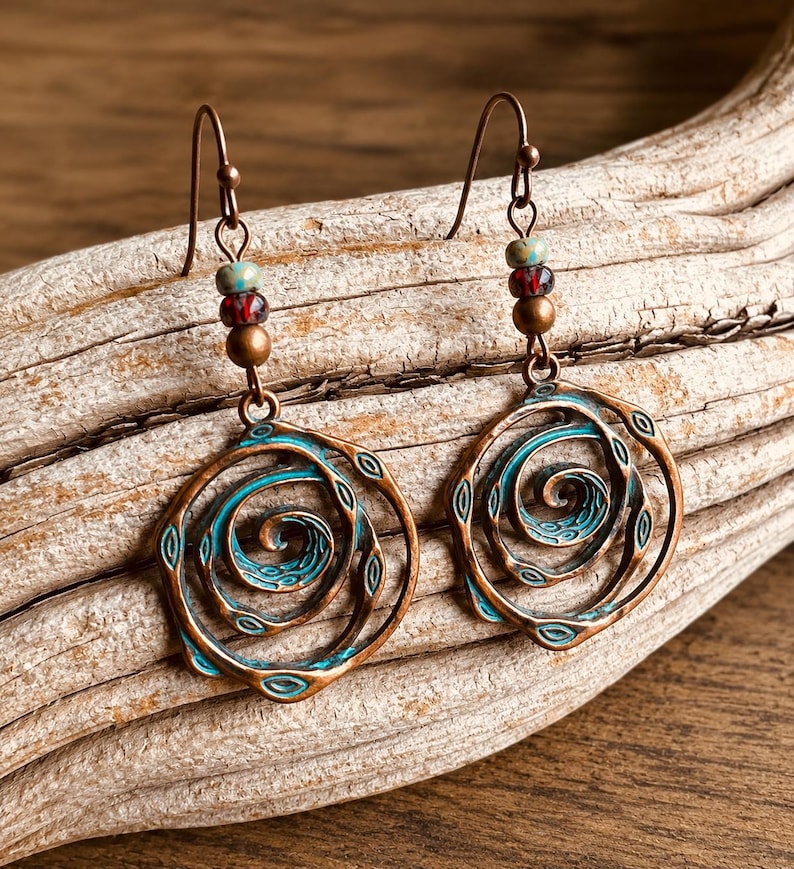 May include: Copper-coloured earrings featuring a spiral design with turquoise detailing. The earrings have a hook fastening and are decorated with small beads in red, green, and brown. The earrings are displayed on a piece of driftwood.
