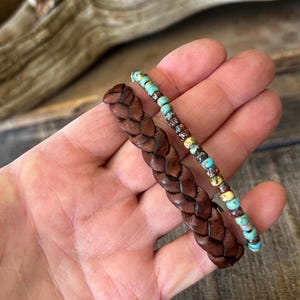 Two bracelets: a braided brown leather bracelet and a beaded bracelet with turquoise, red, and yellow beads. The bracelets are displayed on a wooden surface.