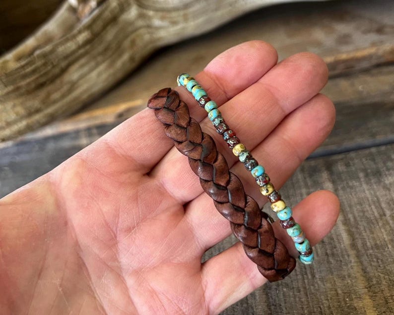 Two bracelets: a braided brown leather bracelet and a beaded bracelet with turquoise, red, and yellow beads. The bracelets are displayed on a wooden surface.