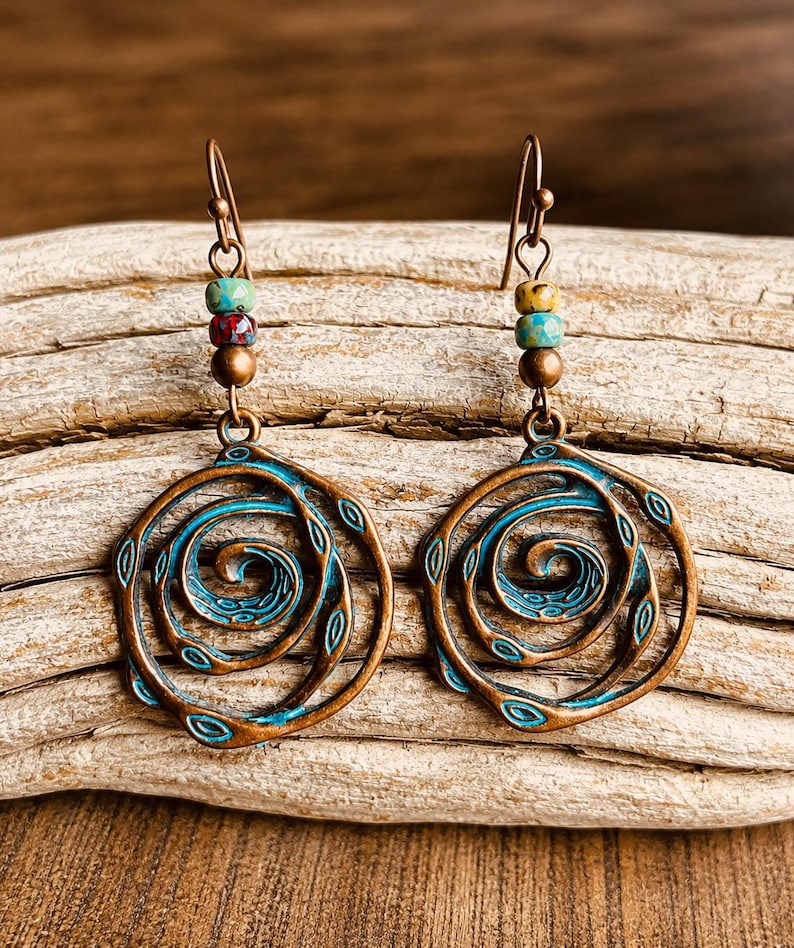 May include: A pair of copper-coloured earrings with a spiral design. The earrings have a turquoise and bronze finish, with colourful beads at the top. The earrings are displayed on a wooden surface.