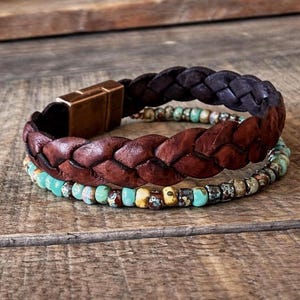 Three bracelets stacked on a wooden surface. The top bracelet is dark brown braided leather with a copper-colored clasp. The middle bracelet is a lighter brown braided leather. The bottom bracelet is made of turquoise and brown beads.