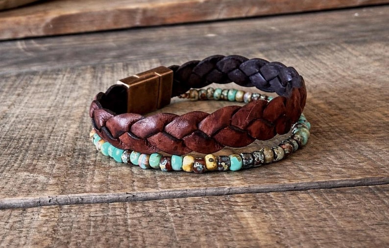 Three bracelets stacked on a wooden surface. The top bracelet is dark brown braided leather with a copper-colored clasp. The middle bracelet is a lighter brown braided leather. The bottom bracelet is made of turquoise and brown beads.