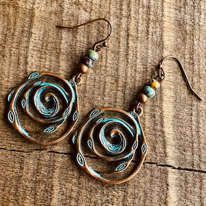 May include: A pair of copper-coloured spiral earrings with turquoise accents and small leaf details. Each earring features a hook and is adorned with colourful beads. The earrings are displayed on a wooden surface.