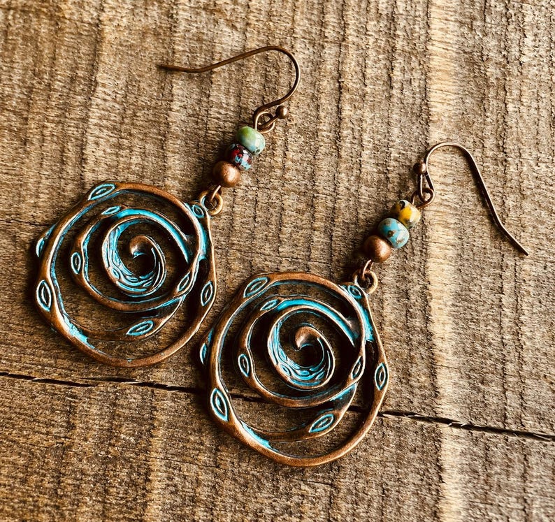 May include: A pair of copper-coloured spiral earrings with turquoise accents and small leaf details. Each earring features a hook and is adorned with colourful beads. The earrings are displayed on a wooden surface.