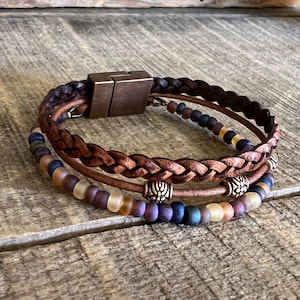 May include: A brown leather braided bracelet with three rows of beads. The beads are in shades of purple, brown, and yellow. The bracelet has a magnetic clasp.