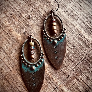 May include: A pair of copper-toned drop earrings featuring a teardrop-shaped metal base with a teal patina finish. Each earring includes a twisted metal oval frame, a central vertical row of three small beads, and a curved ear wire hook.