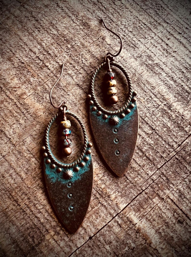 May include: A pair of copper-toned drop earrings featuring a teardrop-shaped metal base with a teal patina finish. Each earring includes a twisted metal oval frame, a central vertical row of three small beads, and a curved ear wire hook.