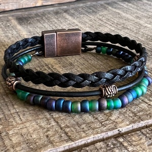 Puede incluir: Un brazalete trenzado de cuero negro con cierre magn&eacute;tico. El brazalete tiene dos filas de cuentas iridiscentes verdes y azules y dos cuentas de metal plateado.