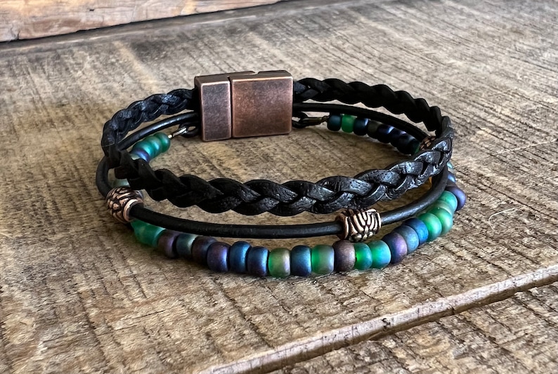 Puede incluir: Un brazalete trenzado de cuero negro con cierre magn&eacute;tico. El brazalete tiene dos filas de cuentas iridiscentes verdes y azules y dos cuentas de metal plateado.