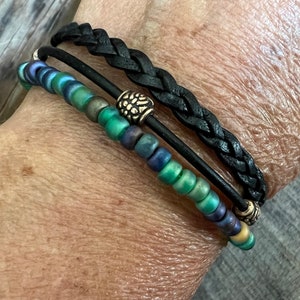 Puede incluir: Un brazalete de capas con una banda de cuero trenzado negro y una hebra de cuentas verdes y azules iridiscentes. El brazalete tiene dos detalles met&aacute;licos plateados.
