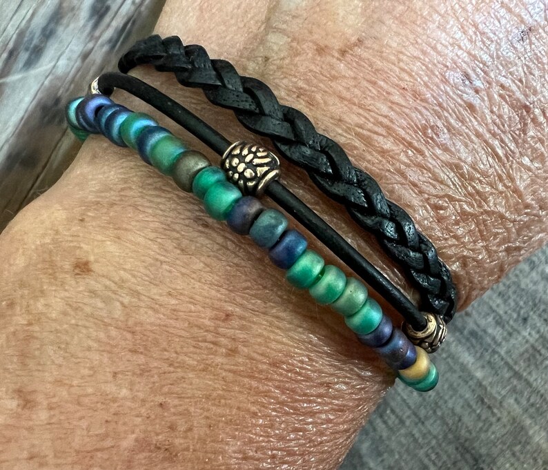 Puede incluir: Un brazalete de capas con una banda de cuero trenzado negro y una hebra de cuentas verdes y azules iridiscentes. El brazalete tiene dos detalles met&aacute;licos plateados.