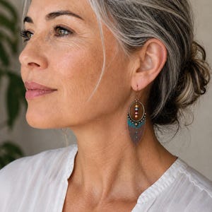 May include: A dangle earring featuring an oval bronze frame with a pointed, leaf-shaped base. The earring includes a vertical row of small, multicolored metallic beads and a textured, teal-colored accent piece at the center of the bronze base.