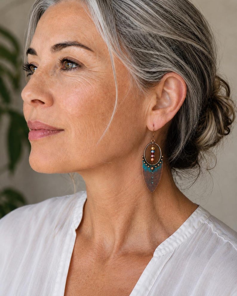May include: A dangle earring featuring an oval bronze frame with a pointed, leaf-shaped base. The earring includes a vertical row of small, multicolored metallic beads and a textured, teal-colored accent piece at the center of the bronze base.