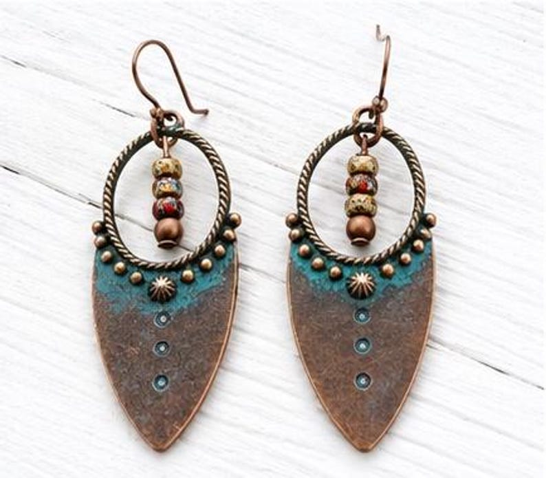 May include: A pair of dangle earrings featuring a copper-toned teardrop-shaped pendant with a teal patina finish, accented by a row of small metal studs and three circular indentations. A twisted metal hoop holds three multi-colored beads.