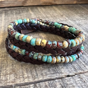 May include: A triple-wrap leather bracelet with a braided leather band and three rows of colorful glass beads. The beads are a mix of turquoise, yellow, and brown.