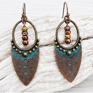 May include: A pair of drop earrings featuring a copper-toned metal shield shape with a teal patina finish. Each earring includes a twisted circular frame, a central star-shaped stud, three small dots, and a vertical row of three multicolored beads.