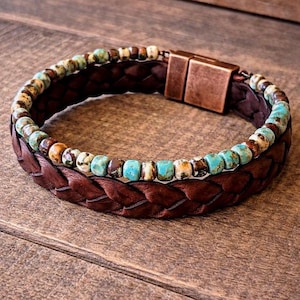 A brown leather bracelet with a braided design and a copper clasp. The bracelet features a strand of turquoise and brown beads. The bracelet is displayed on a wooden surface.