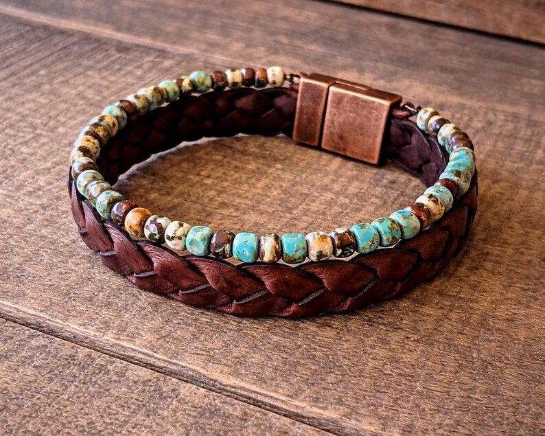 A brown leather bracelet with a braided design and a copper clasp. The bracelet features a strand of turquoise and brown beads. The bracelet is displayed on a wooden surface.