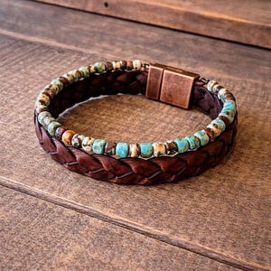 Braided brown leather bracelet with turquoise and brown beads. The bracelet features a copper-colored clasp. The jewelry is displayed on a wooden surface.