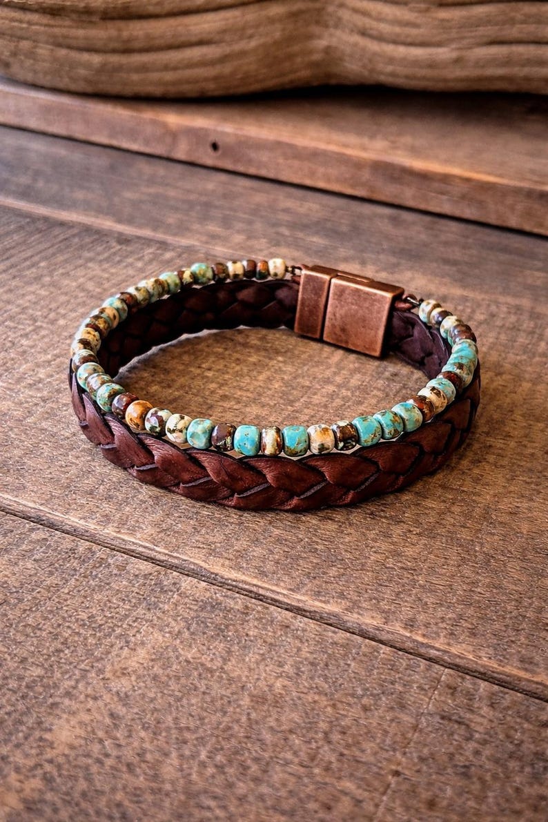 Braided brown leather bracelet with turquoise and brown beads. The bracelet features a copper-colored clasp. The jewelry is displayed on a wooden surface.